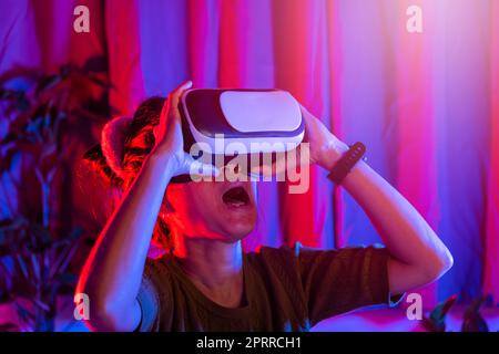 Asiatische Frau tragen den VR-Helm aufgeregt emotional zu Hause lila und blauen Hintergrund, weiblich erleben virtuelle Realität Brille erleben, Stockfoto