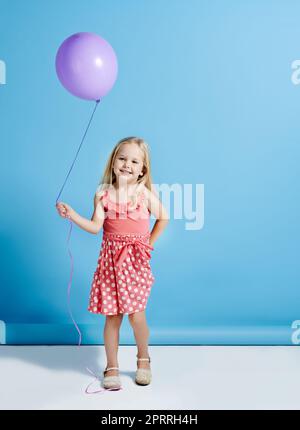 Niemand kann mit einem Ballon entjubelt werden. Ein niedliches kleines Mädchen hält einen Ballon über einem blauen Hintergrund. Stockfoto