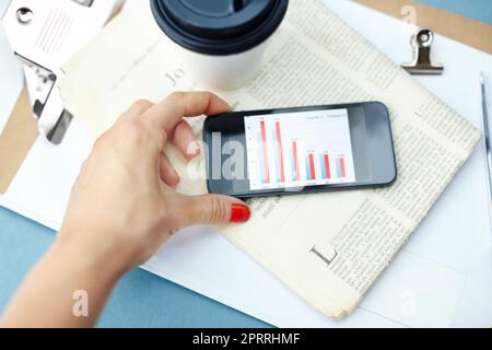 Über aktuelle Ereignisse und Geschäfte auf dem Laufenden bleiben. Handgehaltenes Smartphone mit Balkendiagramm auf dem Bildschirm mit Einwegbecher, Stift, Heftklammer, Zeitung und Klemmbrett. Stockfoto