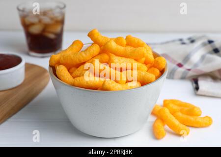 Knuspriger käsiger Maissnack in der Schüssel, Ketchup und erfrischendes Getränk auf weißem Holztisch, Nahaufnahme Stockfoto