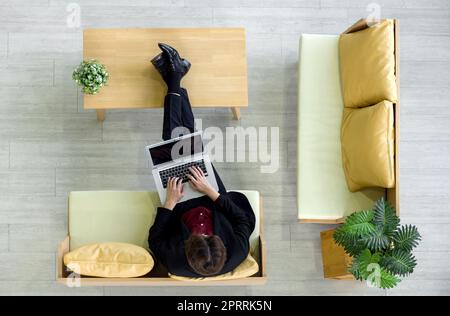 Der junge Geschäftsmann im Anzug sitzt entspannt auf dem Sofa, ruht seine Füße auf dem Tisch, während er auf der Tastatur des Laptops tippt. Draufsicht Stockfoto