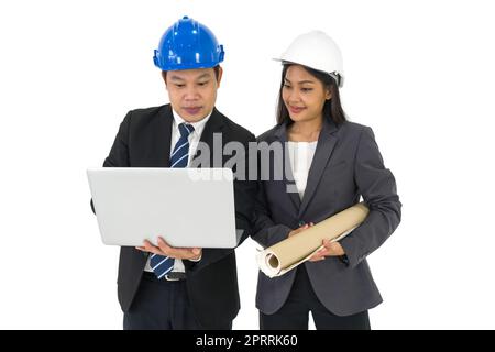 Asiatischer Geschäftsmann in schwarzem Anzug mit blauem Schutzhelm, der auf der Tastatur eines Laptops tippt. Junger Assistent mit Bauzeichnungen von Immobilienprojekten, steht neben ihm. Stockfoto