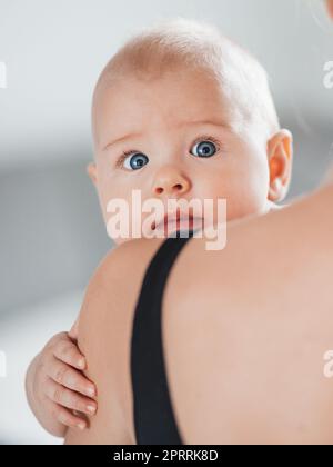 Porträt eines süßen Babys, das in den Armen der Mutter ruht, in die Kamera schaut, die Mutterschulter berührt. Neue mutter hält ein kleines Kind, umarmt das Kind mit Zärtlichkeit, Liebe, Fürsorge. Mutterschaftskonzept Stockfoto
