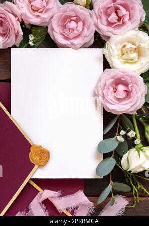 Karte und Umschlag zwischen pinkfarbenen und cremefarbenen Rosen auf braunem Holz mit Blick von oben, Hochzeitsmodell Stockfoto