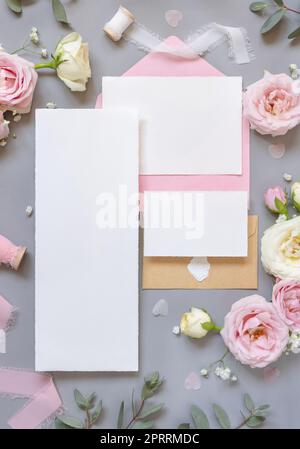 Leere Papierkarten und Briefumschläge zwischen rosa Rosen auf grauer Draufsicht, Hochzeitsmodell Stockfoto