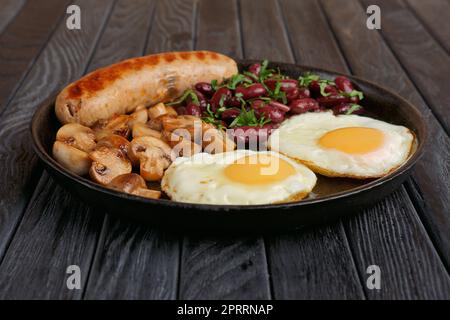 Gusseiserne Bratpfanne mit Spiegeleiern, Würstchen, Pilzen und Bohnen auf einem Holztisch. Ansicht aus niedrigem Winkel. Stockfoto