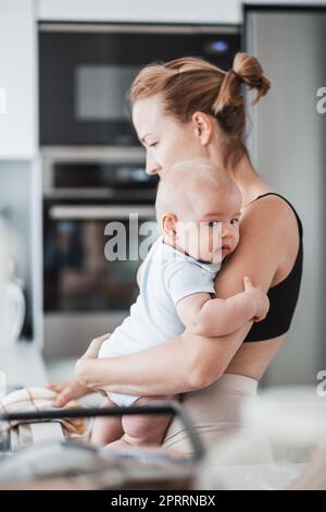 Frau wischt nach dem Abwaschen das Spülbecken mit einem Tuch ab, während sie den vier Monate alten Jungen in den Händen hält Stockfoto