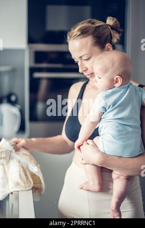 Frau wischt nach dem Abwaschen das Spülbecken mit einem Tuch ab, während sie den vier Monate alten Jungen in den Händen hält Stockfoto