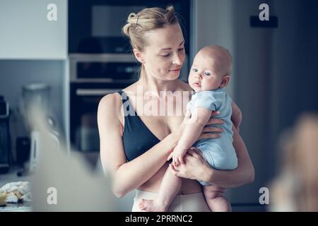 Hübsche junge Mutter hält ihren neugeborenen Jungen zu Hause neben dem Küchenfenster Stockfoto