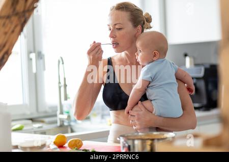 Frau kocht und tast Lebensmittel, während sie ihren vier Monate alten Jungen in den Händen hält Stockfoto