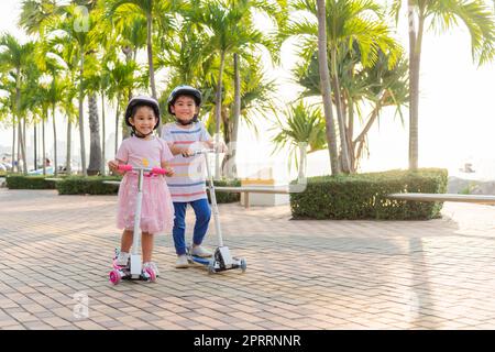 Glückliche asiatische kleine Jungen und Mädchen tragen einen sicheren Helm und spielen Kickboard auf der Straße im Park draußen am Sommertag Stockfoto