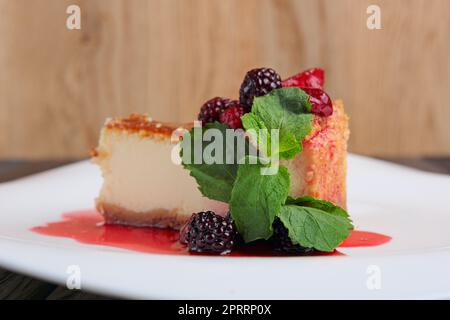 Ein Stück Käsekuchen mit Kirsche, Erdbeere und Taubeere, dekoriert mit Sirup und Minzblättern auf dem Tisch Stockfoto