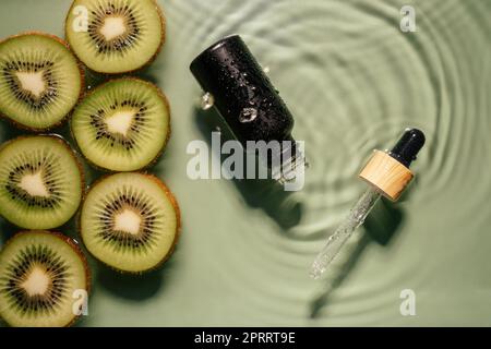 Gesichtspipette Serum schwarze Flasche, Molke Beauty-Produkt in der Nähe von Obst tropischen Kiwi Scheiben in Wasser spritzt frische transparente, Flecken. Draufsicht. Organisch Stockfoto