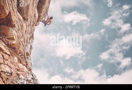 Klettern, Sport und Abenteuer mit einem Mann auf dem Berg vor blauem Himmel für Fitness, Herausforderung und Training. Motivation, Freiheit und starker männlicher Athlet auf der Klippe für Adrenalin Stockfoto