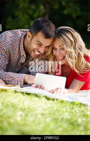 Auf keinen Fall. Ein lachendes junges Paar, das ein digitales Tablet benutzt, während es auf dem Gras in einem Park liegt. Stockfoto