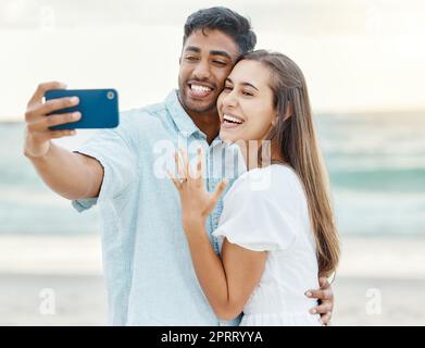 Liebe, Strand und Paar nach ihrer Verlobung machen ein Selfie am Telefon während des Sommerurlaubs. Glückliche Frau, die nach einem Heiratsantrag ihren Ring vorführt, während sie im Urlaub mit einem Mann fotografiert. Stockfoto