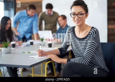 Sie sticht im Büro hervor. Porträt einer attraktiven jungen Frau, die in einem Büro mit Kollegen im Hintergrund sitzt. Stockfoto