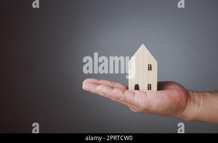 Konzept des Verkaufs eines Hauses. Eine Hand hält ein Modellhaus auf grauem Hintergrund. Immobilienmakler bieten Haus, Sachversicherung und Sicherheit, erschwingliche Wohnkonzepte, Hausversicherungsvermittler, Verkäufer. Stockfoto