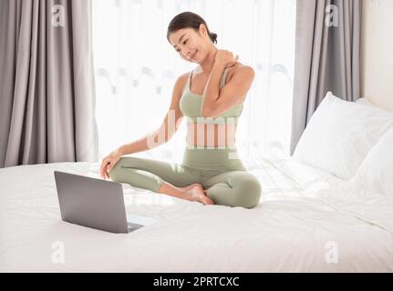 Frau, die Yoga im Bett praktiziert, nachdem sie aufgewacht ist. Asiatische Frau, die auf dem Bett sitzt und ihren Laptop benutzt, während sie zu Hause Dehnübungen macht. Stockfoto