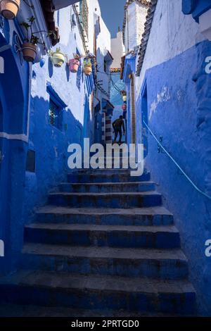 Silhouette eines Mannes, der die Treppe einer steilen Gasse in der blauen Medina von Chefchaouen hochklettert, dekoriert mit Töpfen in verschiedenen Farben. Stockfoto