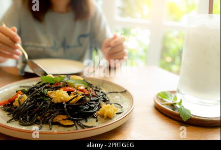 Schwarze Tintenfisch-Tintenfisch-Spaghetti mit Garnelen auf dem Teller. Schwarze Pasta mit Tintenfischtinte auf einem Restauranttisch und verschwommene Frau, die mit Gabel und Löffel isst. Gesundes Essen. Eine Frau isst köstliche schwarze Spaghetti. Stockfoto