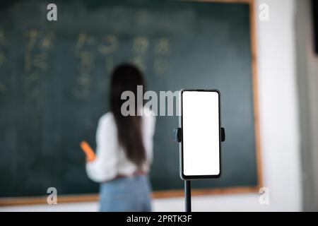 Asiatische Lehrer unterrichten an Bord, während sie online über Smartphone im Klassenzimmer Tafel-Vorlesungen halten Stockfoto