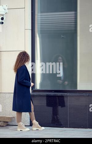 Nicht erkennbare Frau im Mantel geht den Bürgersteig entlang und schaut auf ihre Spiegelung in den Fenstern Stockfoto