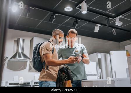 Verkäufer und reifer Mann diskutieren über Smartphone im Haushaltswarenladen Stockfoto