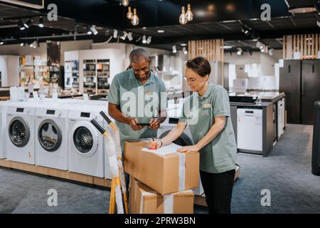 Lächelnder Verkäufer mit Taschenrechner, während eine Verkäuferin ein Dokument auf einem Karton im Haushaltswarenladen schreibt Stockfoto