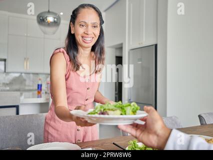 Ehefrau, gesundes Essen und Salat, während sie Lunch oder Dinner für Ehemann mit einem Lächeln zu Hause servieren. Eine fürsorgliche und glückliche Hausfrau mit einem Essteller und einer veganen Mahlzeit am Tisch Stockfoto