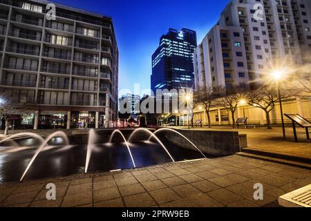 Die Sonne geht über der Stadt unter. Ein Brunnen, der nachts auf einem leeren Stadtplatz fließt. Stockfoto