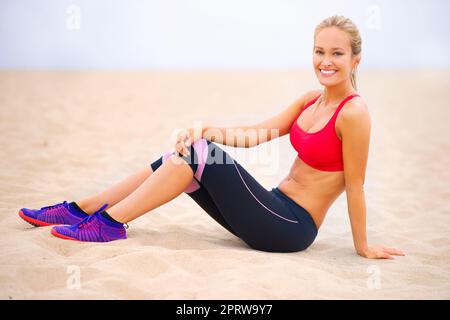 Getrimmt und getönt. Porträt einer jungen Frau in Sportkleidung, die am Strand sitzt. Stockfoto
