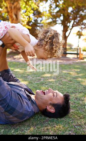 Familienlachen ist ansteckend: Ein Vater liegt auf dem Gras, während er seinen Teigschaber hebt. Stockfoto