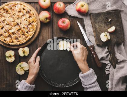 Eine Frau legt Apfelscheiben in ein rundes Backblech auf den Tisch, neben die Zutaten Stockfoto