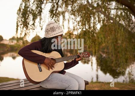 Sie kommt hierher, um Inspiration zu finden. Eine schöne junge Frau, die Gitarre spielt, während sie auf einer Bank neben einem See sitzt. Stockfoto