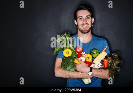 Essen Sie gut, sehen Sie gut aus, fühlen Sie sich gut. Studioporträt eines jungen Mannes, der eine Armvoll gesundes Gemüse vor dunklem Hintergrund trägt Stockfoto