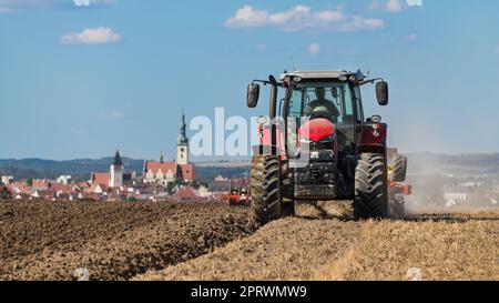 Roter Traktor auf dem Feld beim Pflügen von Stoppeln und grauenhaftem Schmutz in tschechischer Landschaft. Funktionierendes Landwirtschaftsfahrzeug mit Blick auf den Kirchturm in der historischen Stadt Tabor. Stockfoto