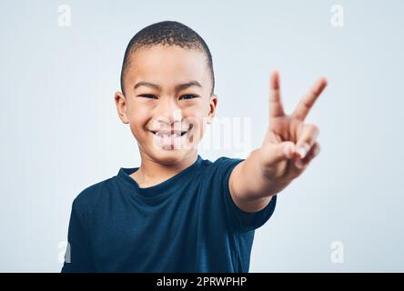 Halten Sie es cool yall. Studioaufnahme eines niedlichen kleinen Jungen, der vor grauem Hintergrund ein Friedenszeichen macht. Stockfoto