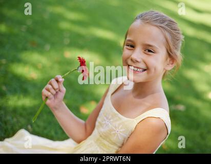 Eine schöne Blume für ein schönes Mädchen. Ein Porträt eines schönen kleinen Mädchens, das draußen auf dem Gras mit einer schönen roten Blume sitzt. Stockfoto