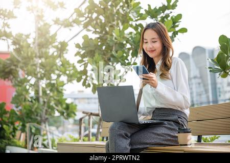 Junge Frau, die mit einem Laptop arbeitet und ein mobiles Smartphone im Außenbereich eines Gebäudes verwendet Stockfoto