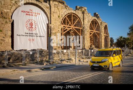 Ein gelbes Taxi fährt an den Ruinen des Amphitheaters in der antiken römischen Stadt Side in der Provinz Antalya, Türkei (Turkiye) vorbei. Das Amphitheater wurde gebaut Stockfoto