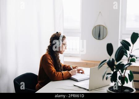 Junger Student, der ein virtuelles Seminar besucht, während er zu Hause studiert. Frau trägt Kopfhörer, schreibt auf dem Laptop und macht sich während des E-Learning Notizen Stockfoto