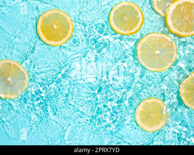 Zitronenscheiben in sauberem, transparentem Wasser auf blauem Hintergrund mit Kopierfläche. Wasser spritzt im Sonnenlicht auf die blaue Wasseroberfläche. Draufsicht oder Flatlay. Stockfoto
