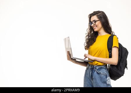 Studentin isoliert auf grauem Hintergrund, lächelt vor der Kamera, drückt den Laptop an die Brust, trägt einen Rucksack, ist bereit zum Lernen, startet einen neuen Profi Stockfoto