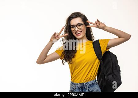 Schöne charismatische kaukasische Rotschopf Mädchen mit Rucksack, Kopfhörer über dem Hals, zeigt Friedenszeichen und lächelnd erfreut und freudige Kamera, stehend wh Stockfoto