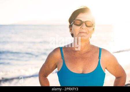 Seniorin am Strand, am Meer. Er trägt blaue Badebekleidung und eine Schwimmbrille, bereit zum Schwimmen Stockfoto