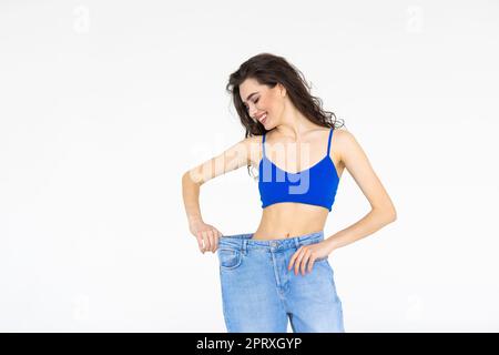 Schlanke junge Frau in alten großen Jeans, zeigt erfolgreiches Ergebnis einer Diät - Gewichtsverlust, schlankes Konzept. Weißer Hintergrund Stockfoto