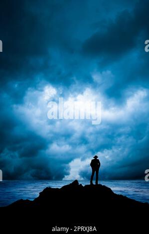 Ein Mann mit einem Fedora-Hut, der auf einem Felsen in der Nähe des Meeres steht Stockfoto