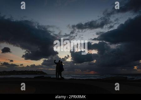 Ein Paar, das den wunderschönen Sonnenuntergang am Strand mit stürmischen Wolken einfängt Stockfoto