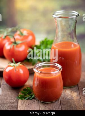 Tomatensaft in Glas und Karaffe mit frischen Tomaten und Petersilie auf Holztisch mit Sommerhintergrund Stockfoto